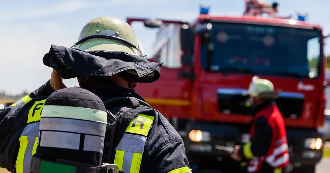 Feuerwehrmann in Uniform und Sauerstoffflasche, im Hintergrund Feuerwehrwagen